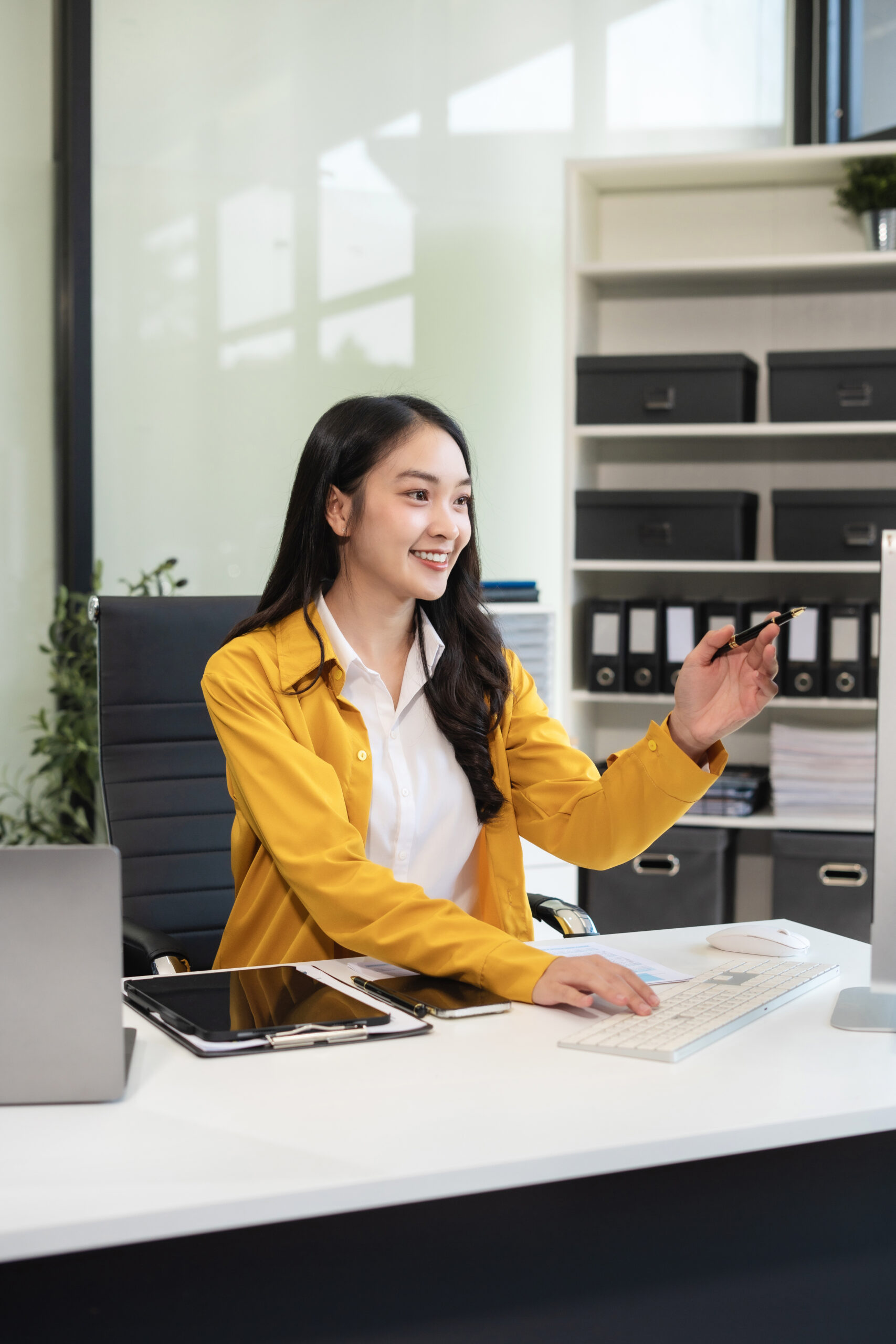 Smiling Asian businesswoman points at computer screen, working on financial and marketing data in a modern office.