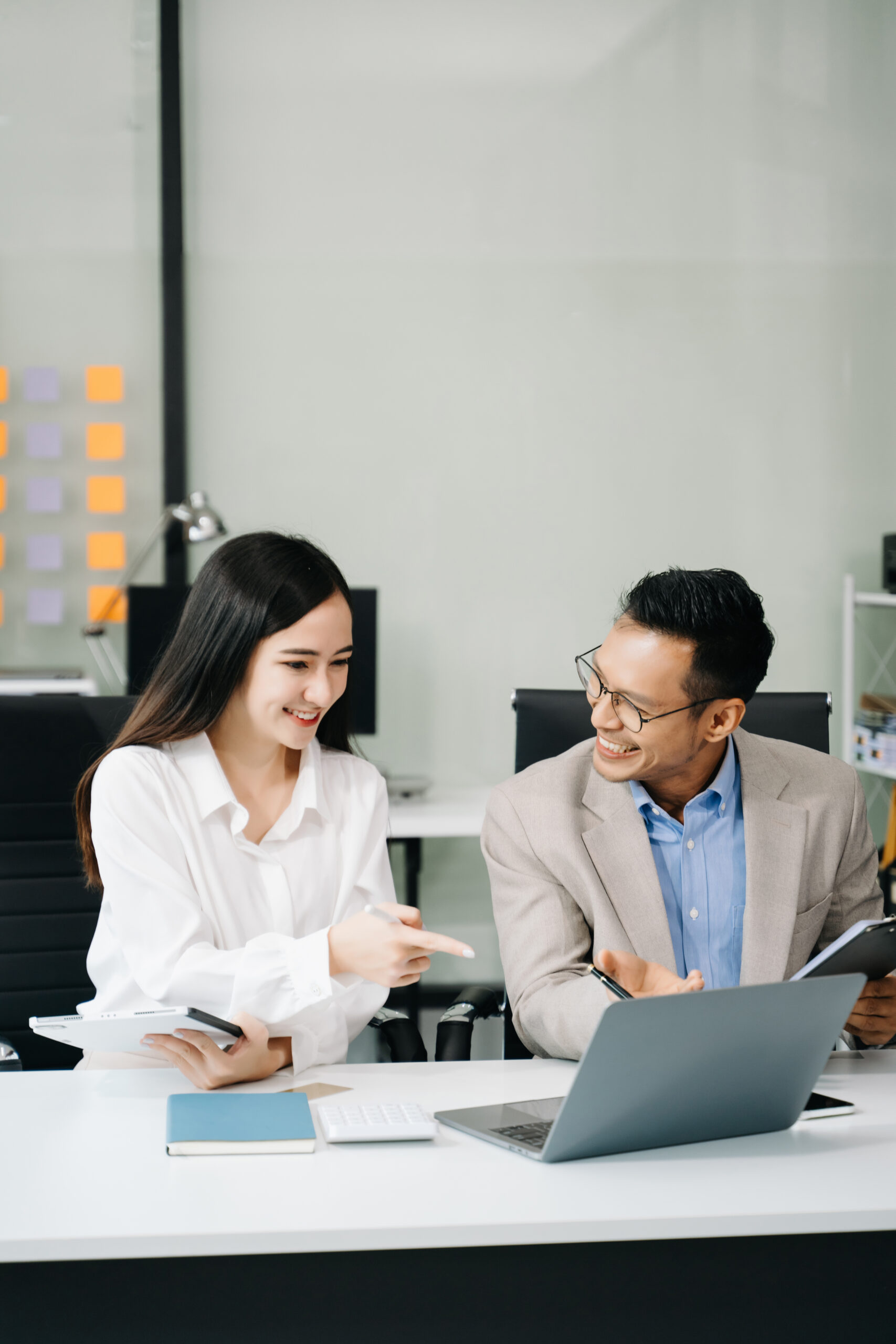 Two smiling Asian business professionals collaborating at a desk with a laptop and tablet, demonstrating efficient teamwork for a multi-channel contact center.
