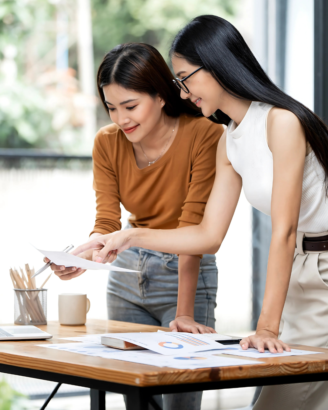 Two happy Asian businesswomen collaboratively reviewing documents and charts, demonstrating efficient coworking for outsourced financial services.